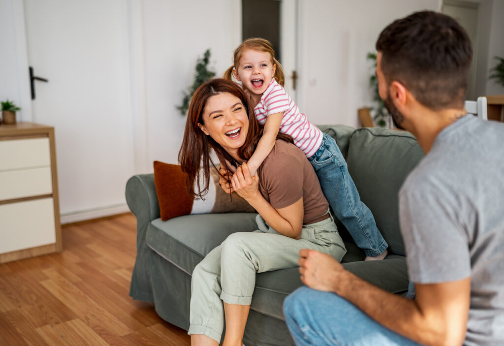 Happy family spending time at home, mother laughing while daughter plays on her back, father watching